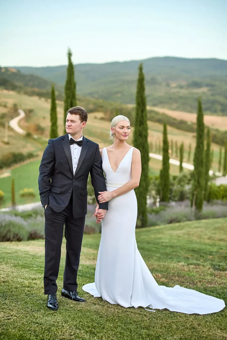 Bride and groom holding hands on a lawn, with cypress trees and rolling Tuscan hills behind them